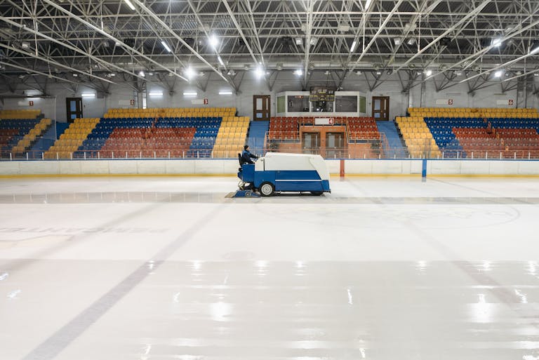 A Zamboni ice resurfacer operating in an empty indoor ice skating rink with colorful seating.
